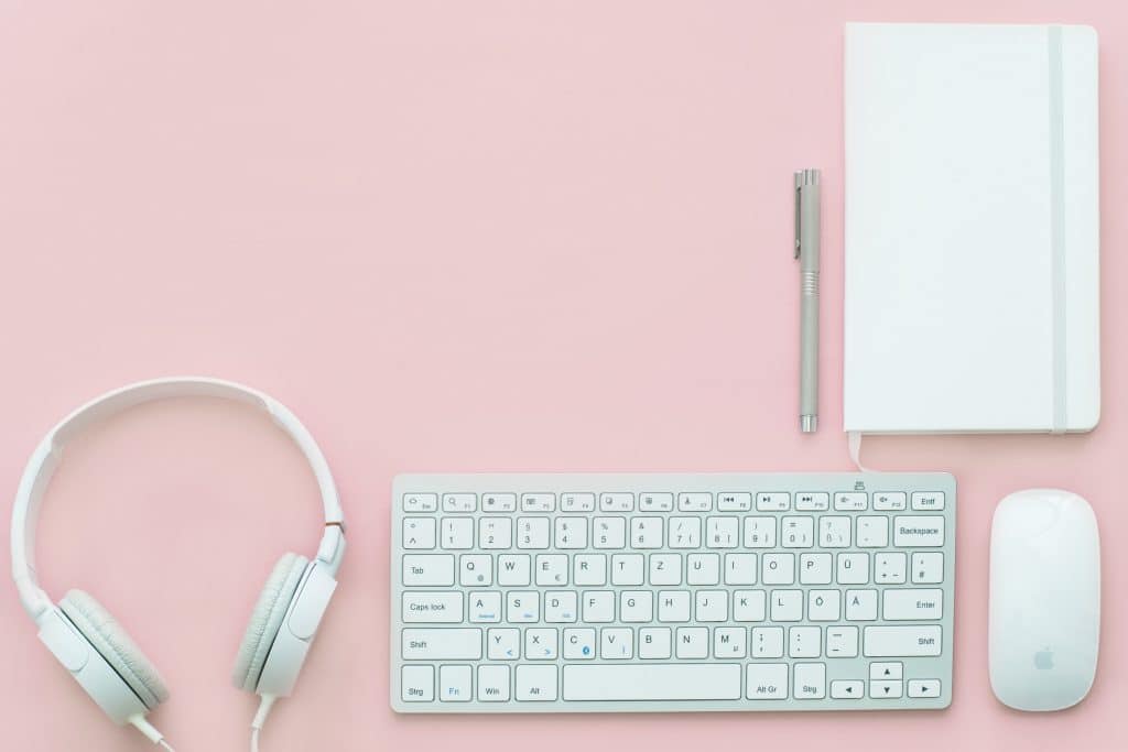 Un teclado de ordenador, un ratón, una libreta con un bolígrafo y unos auriculares sobre un fondo rosa son parte de mis herramientas para lograr un diseño web personalizado - Maria en la red